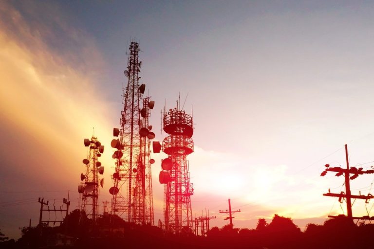Cluster of tall communication towers with satellite dishes silhouetted against a sunset sky with an orange glow on the horizon.