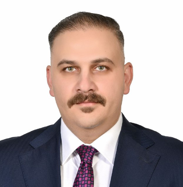Professional headshot of a man with a mustache in a navy suit and pink patterned tie against a white background, shoulders visible.