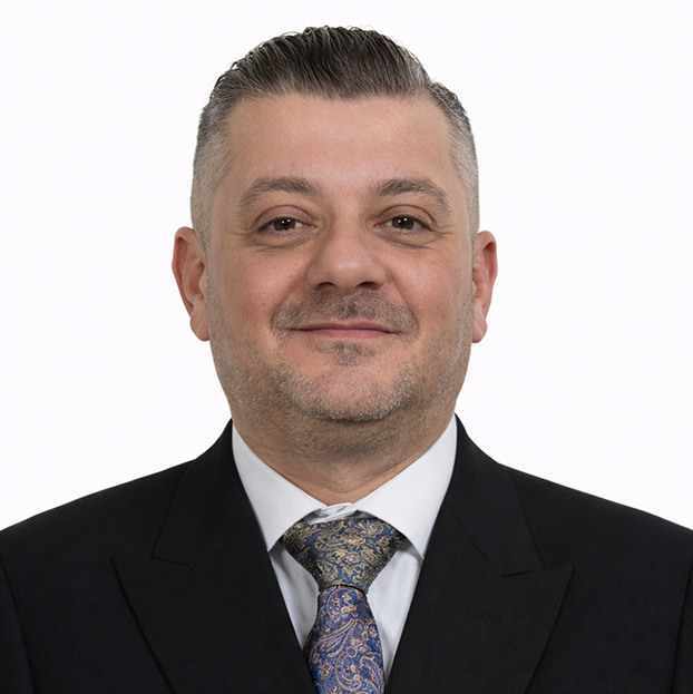 Portrait of a smiling man in a black suit, white shirt, and paisley tie against a white background.