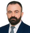Portrait of a man with dark hair and a full beard, wearing a navy suit and tie, looking at the camera against a light background.