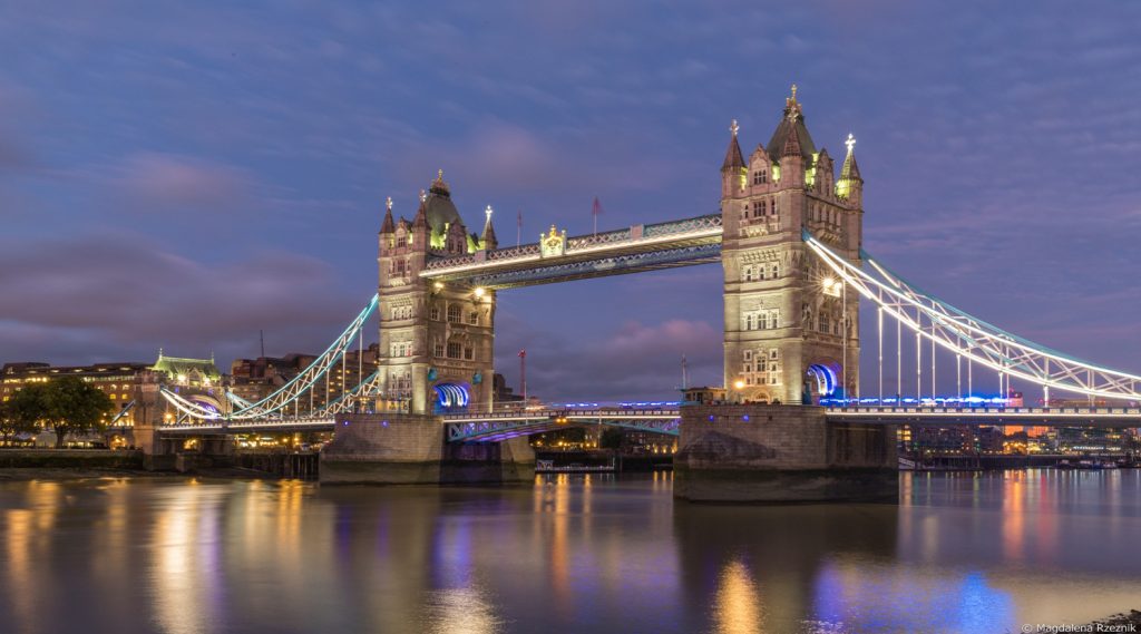 Tower Bridge illuminated at dusk over the River Thames, with reflections on calm water.”] ,