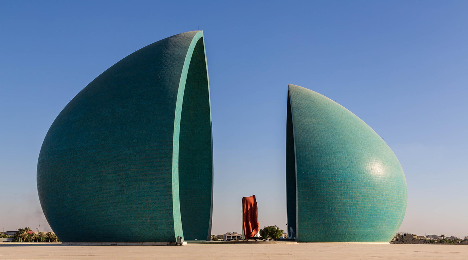 Two large green sail‑shaped towers split by a central gap, with a red sculpture between them on a wide plaza under a blue sky.