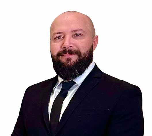 Professional headshot of a bald man with a beard wearing a black suit and tie, looking at the camera against a white background.
