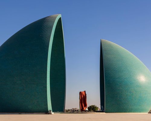 Two large green sail‑shaped towers split by a central gap, with a red sculpture between them on a wide plaza under a blue sky.