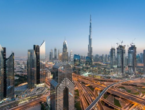 Panoramic view of Dubai's skyline with the Burj Khalifa and modern high-rises, plus intricate highway interchanges below at dusk/dawn.